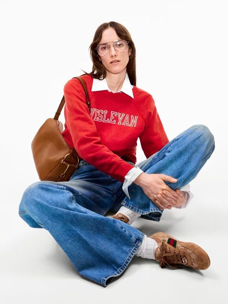 A woman in a red sweater, wide-leg jeans, and sneakers sits cross-legged against a white backdrop, carrying a brown leather shoulder bag.