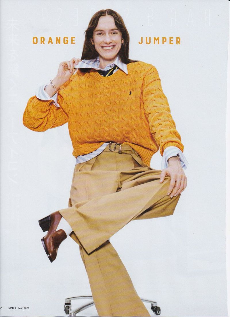 Smiling woman in an orange cable-knit sweater, light-blue collared shirt, and khaki pants seated on a rolling chair against white backdrop.