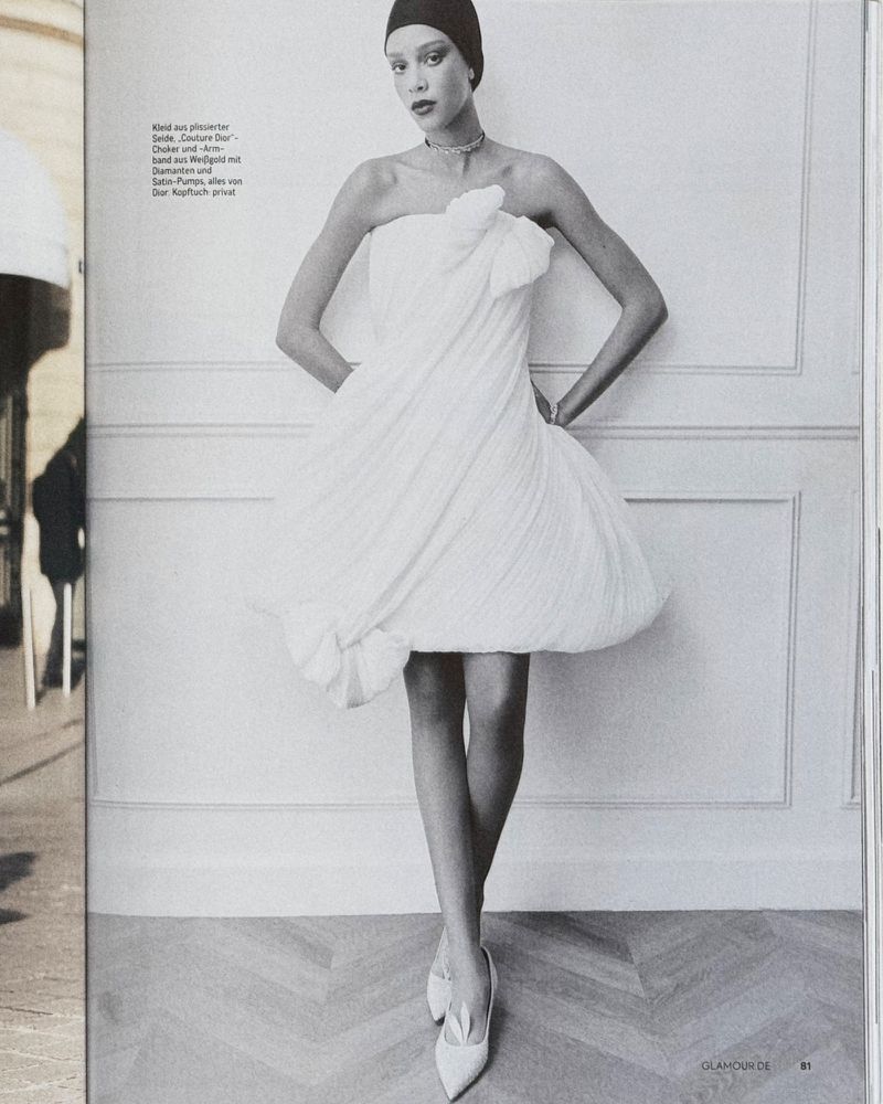 Glamour fashion shot: A woman in a white strapless dress with a black cap poses indoors against a neutral wall and parquet floor.