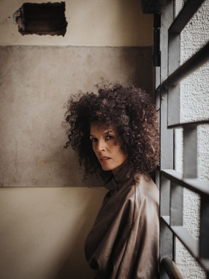 A woman with curly hair stands in front of a window with bars in an indoor setting, wearing a brown jacket.