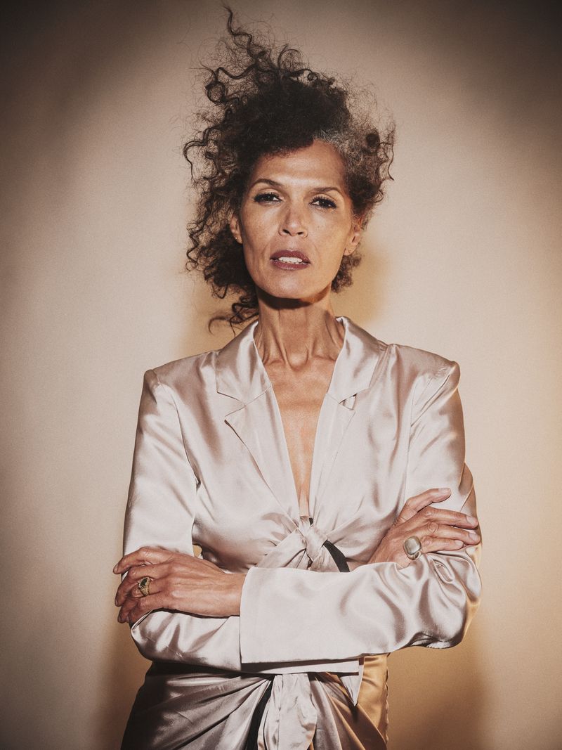 Portrait of an adult woman with curly hair, arms crossed, wearing a silky jacket and skirt, set against a neutral background.