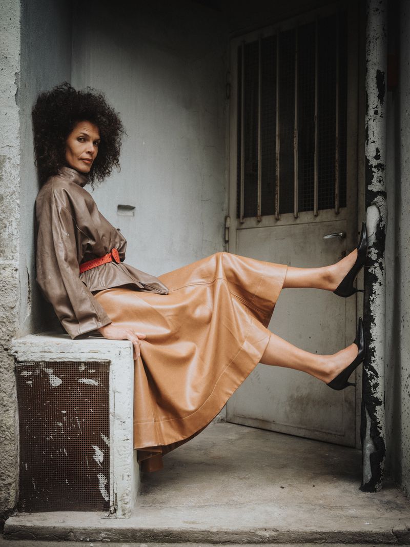 A woman in a brown leather jacket and skirt sits in front of a metal door and weathered wall, wearing black high heel shoes.