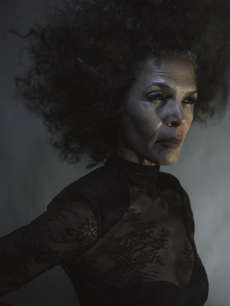 Portrait of a mature African American woman with an afro hairstyle wearing a black lace top, shot against a dark, smoky background.