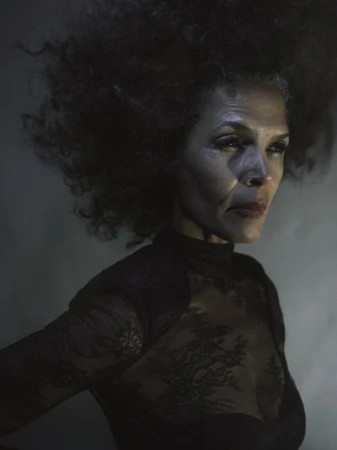Portrait of a mature African American woman with an afro hairstyle wearing a black lace top, shot against a dark, smoky background.