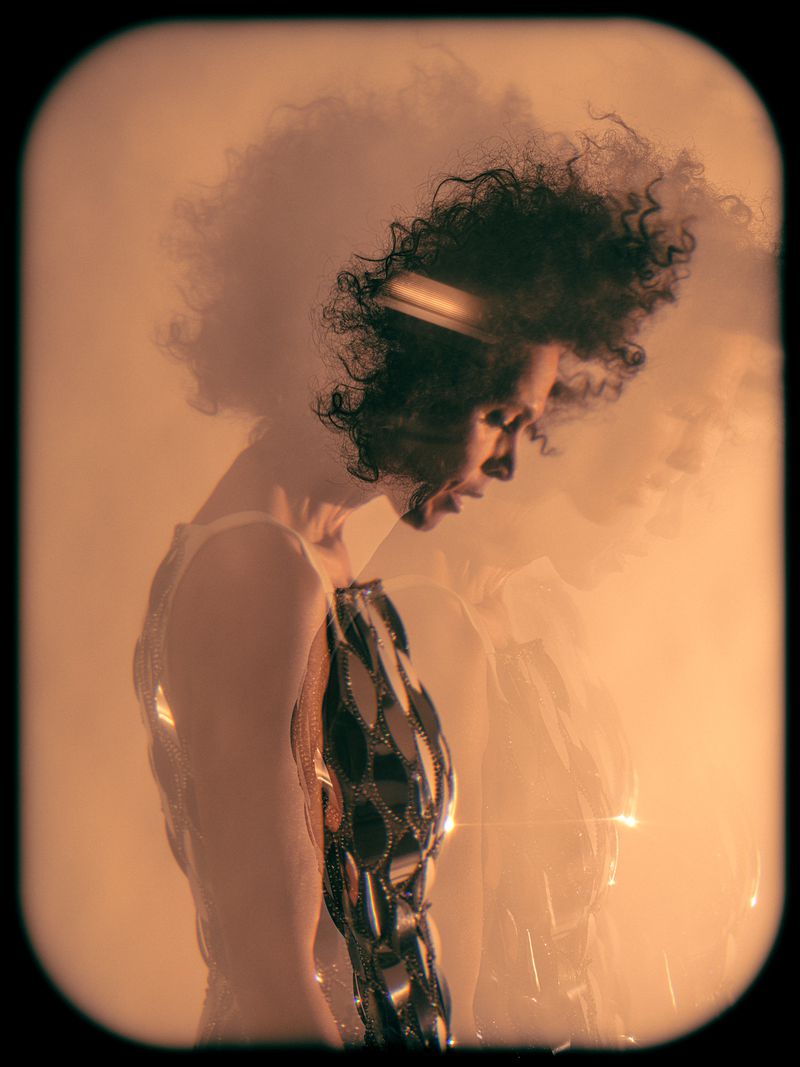 Side profile studio shot of a woman with dark curly hair, wearing a sleeveless silver dress with mirrored accents against an orange gradient background.