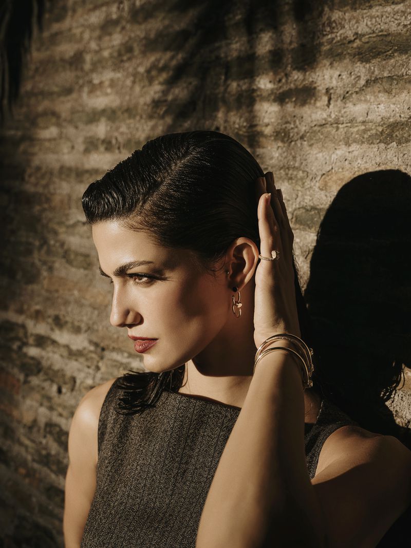 Elegant adult woman with hand to her head, accessorized with bracelets and earrings, illuminated by warm lighting against a textured wall.