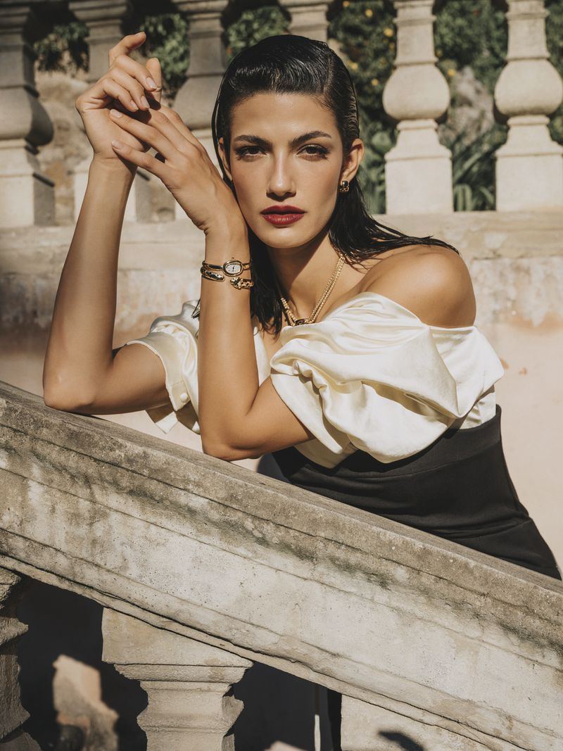 Woman in elegant off-shoulder dress poses on stone steps with jewelry, dark hair, and red lipstick.