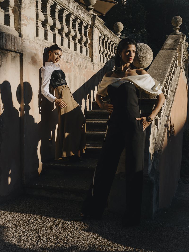 Two adult women in fashionable jumpsuits pose on stone steps with an ornate stone railing in a sunlit outdoor setting.
