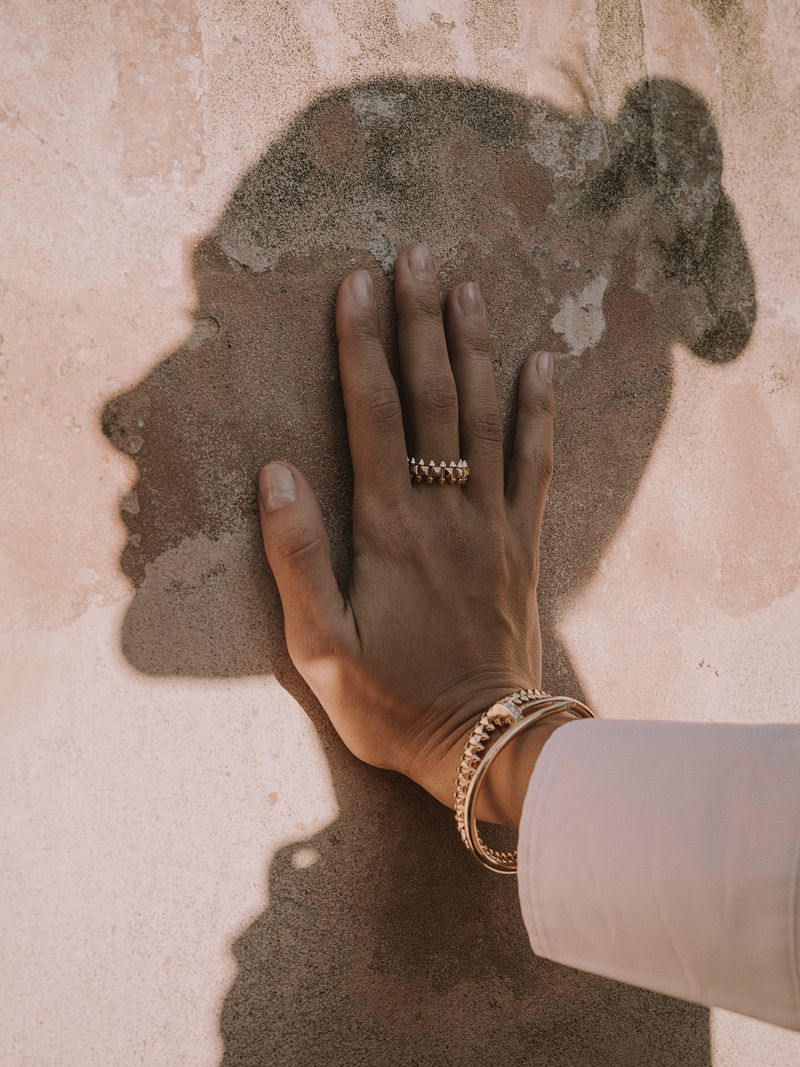 Close-up of a person's hand wearing a gold ring and bracelets, casting a shadow of a face against a textured peach-toned wall.