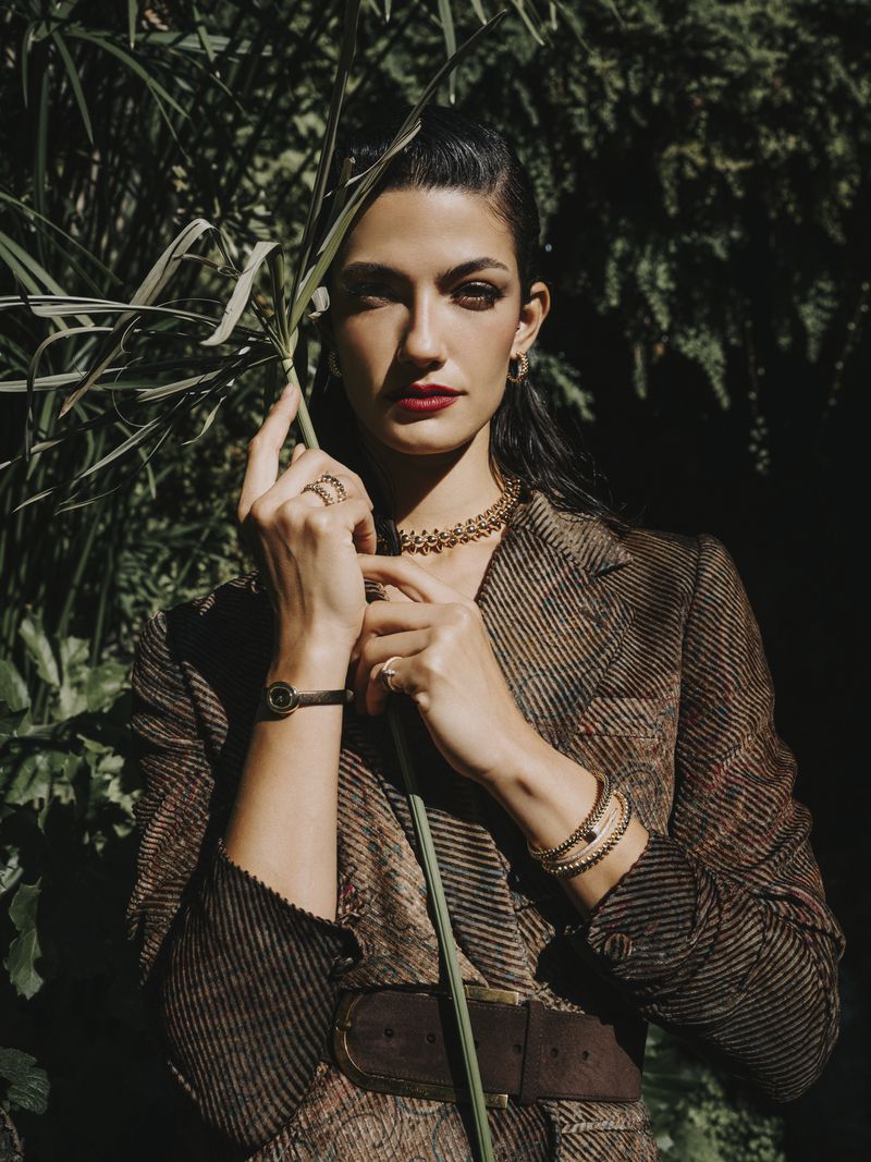 Elegant woman with dark hair and red lipstick poses in a brown herringbone blazer, accessorized with jewelry, amid leafy green foliage.