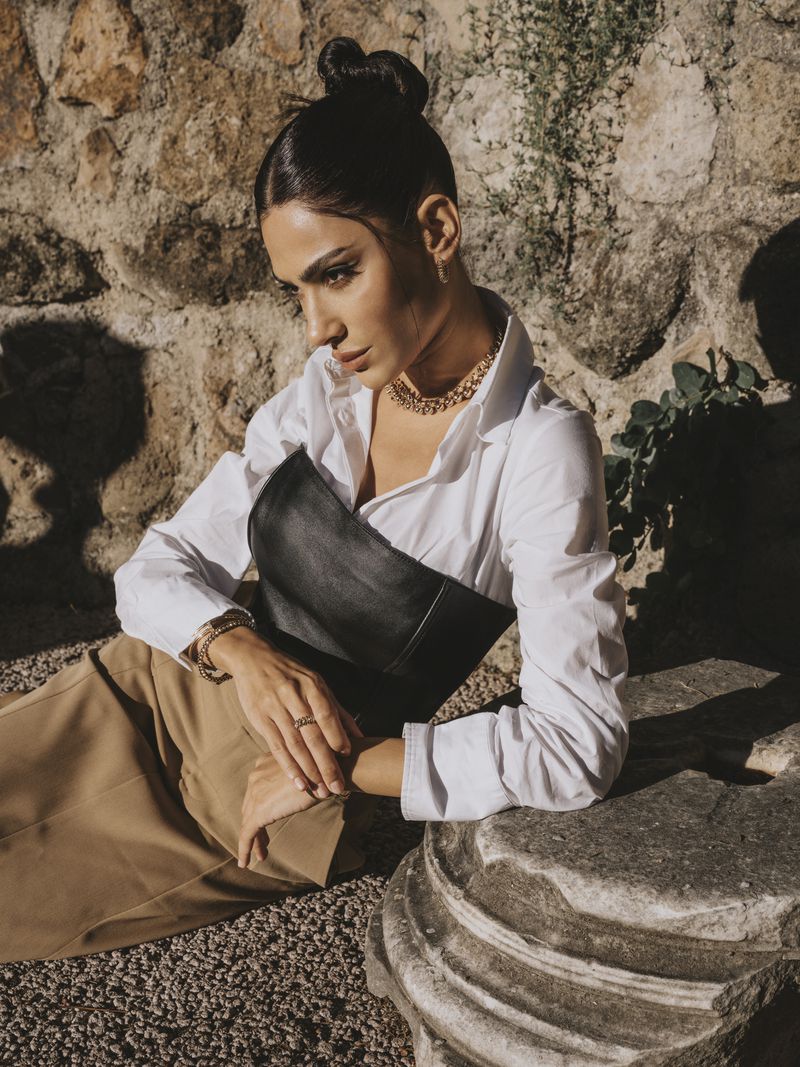 Portrait of a beautiful adult woman in a white blouse and black corset posing against a stone wall with natural light and tan gravel.