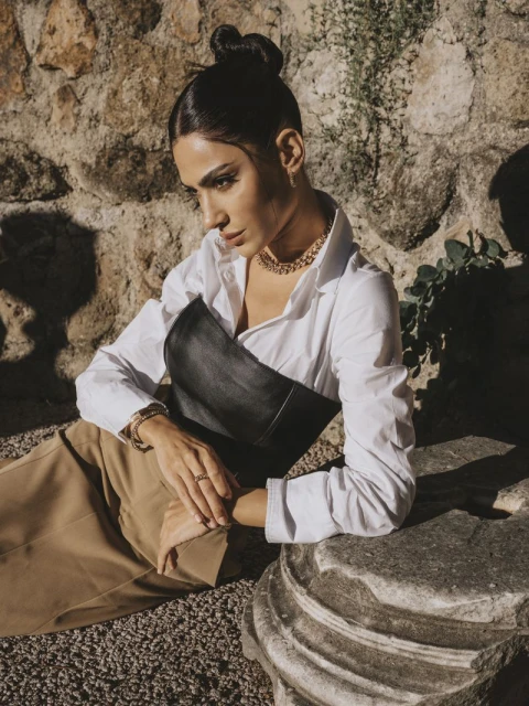 Portrait of a beautiful adult woman in a white blouse and black corset posing against a stone wall with natural light and tan gravel.