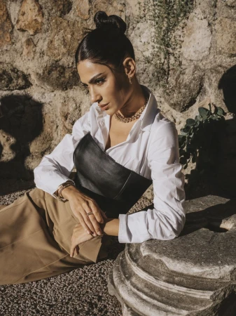 Portrait of a beautiful adult woman in a white blouse and black corset posing against a stone wall with natural light and tan gravel.