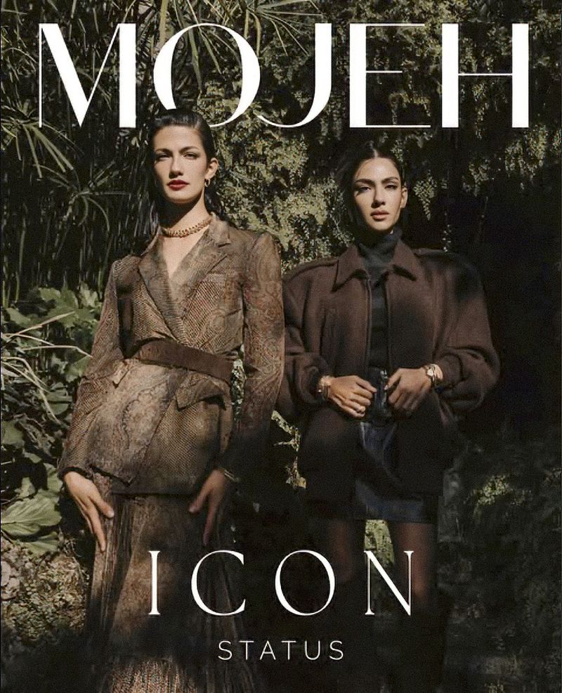 Two fashion models pose in a leafy outdoor setting for a magazine cover, wearing stylish brown blazers, skirts, and turtleneck tops.