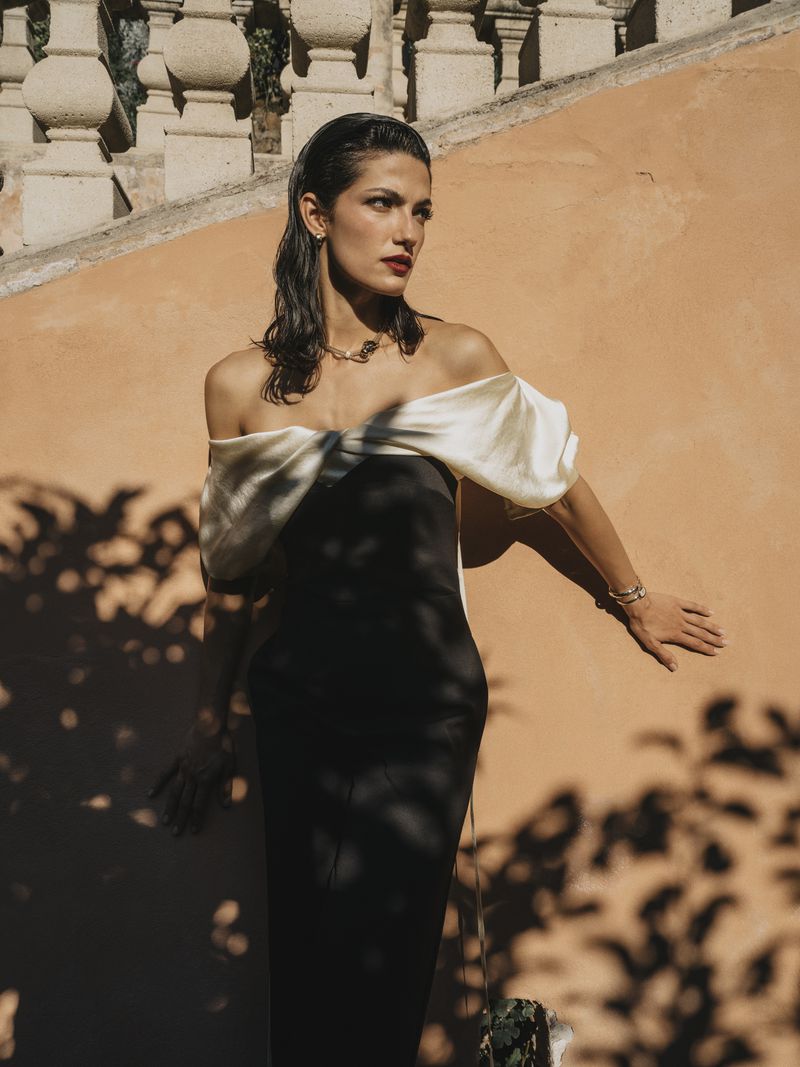 Huda El Mufti posing in an off-the-shoulder black dress with white draped satin neckline against a terracotta wall with leafy shadows.