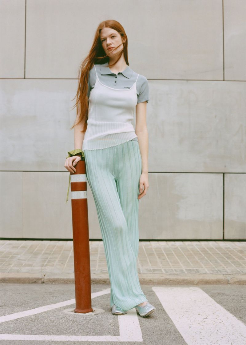 Red-haired woman poses in pleated green pants and top against a textured grey wall, with a red and white striped bollard nearby.