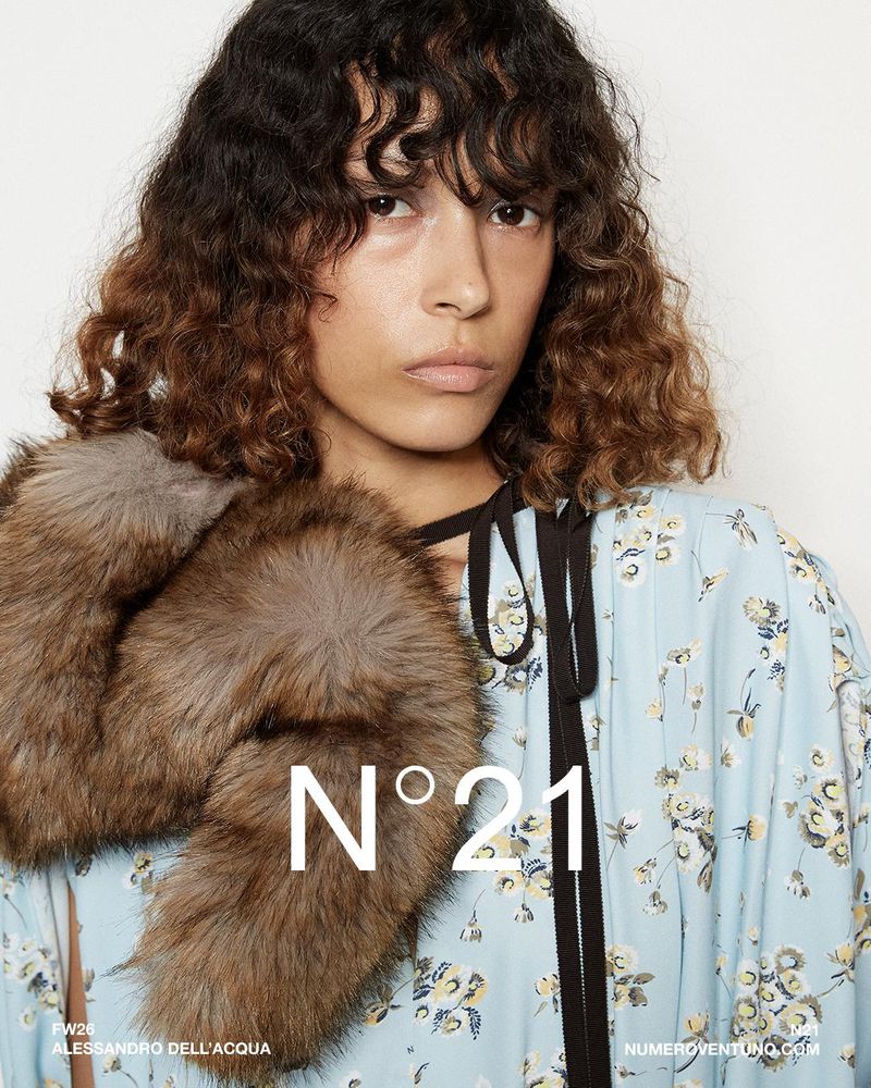 Fashion shot of a woman wearing a floral dress with a fur stole, posing in a studio. The brand logo is visible on the image.