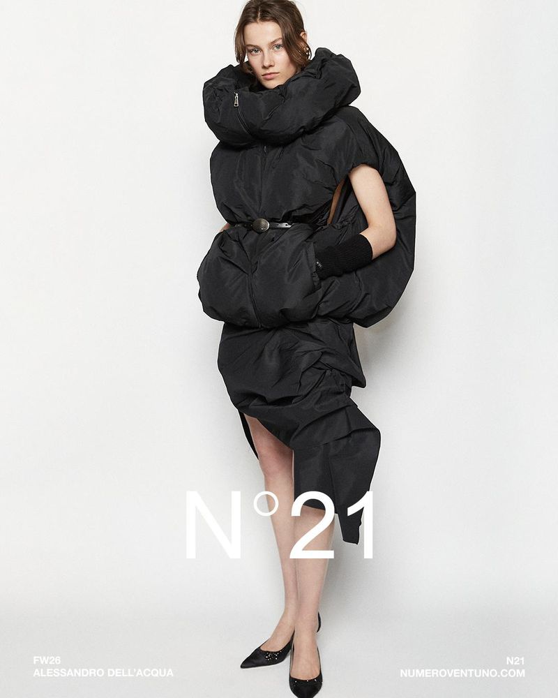 Fashionable woman poses in black oversized puffer coat and heels against white backdrop, showcasing modern, avant-garde style and design.
