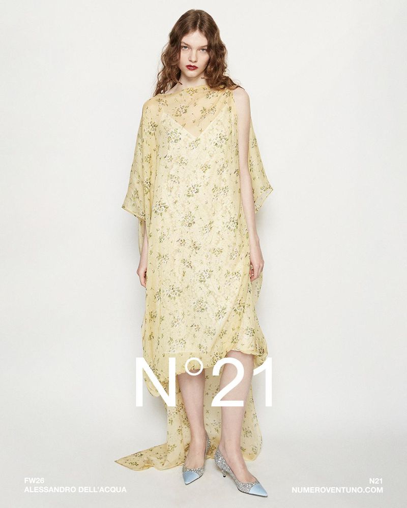Fashion shot of a female model wearing a yellow floral gown with a sheer overlay, accessorized with light blue heels against a white backdrop.