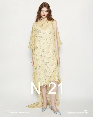 Fashion shot of a female model wearing a yellow floral gown with a sheer overlay, accessorized with light blue heels against a white backdrop.