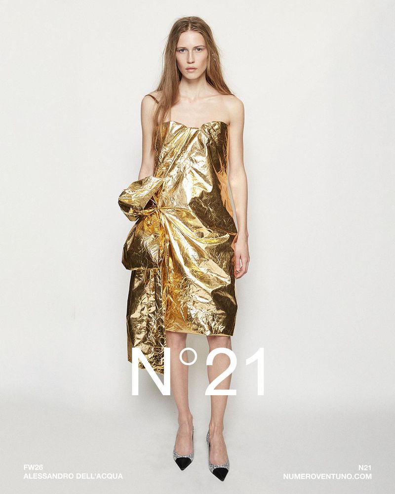A fashion model wearing a metallic gold strapless dress with draped fabric at the waist against a plain white background.