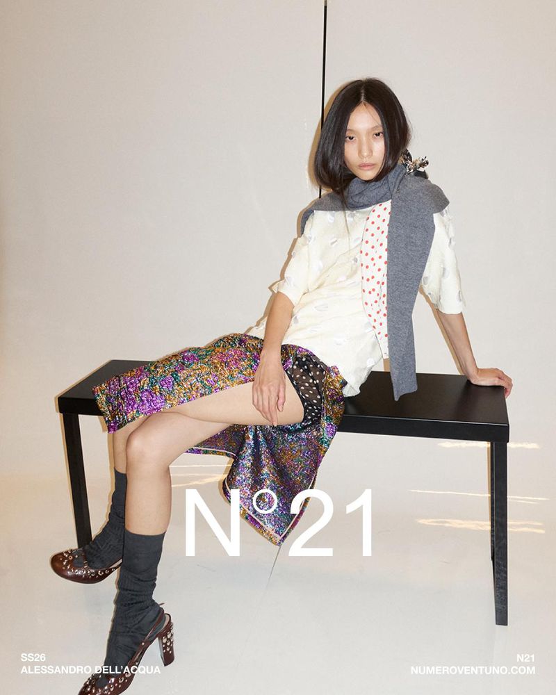 Asian woman poses on a black bench wearing a white shirt, grey scarf, and multi-colored sequin skirt with black heeled Mary Janes in a studio.