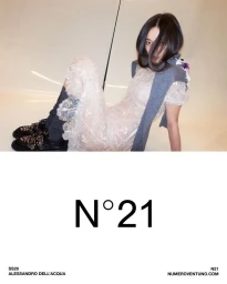 Model in a sheer white lace dress and grey sweater seated against a neutral studio backdrop for N21 SS26 campaign.