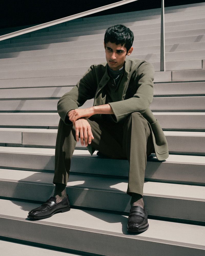 Man in olive green suit sitting on steps with black loafers. Dark hair, light skin, hands clasped in casual pose. Neutral background.
