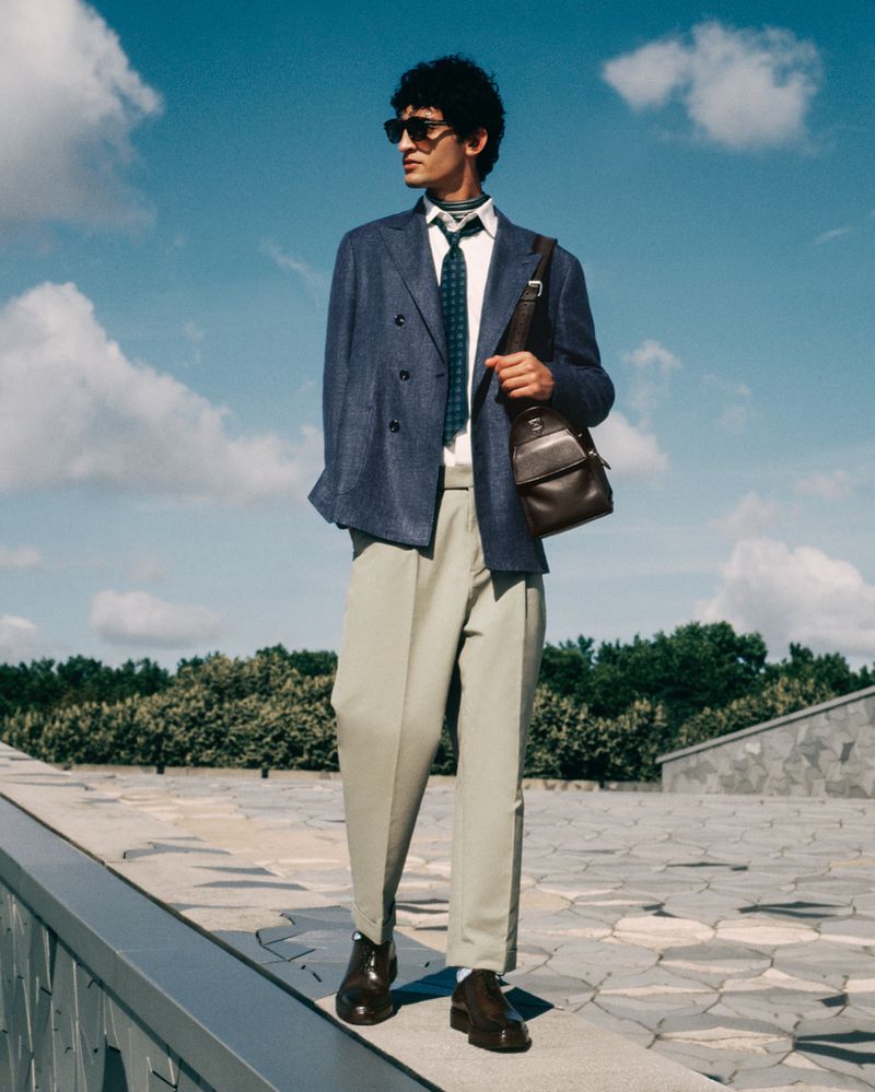 Man in a double-breasted blue blazer, tan pants, and sunglasses stands outdoors with a brown leather messenger bag and a blue patterned tie.