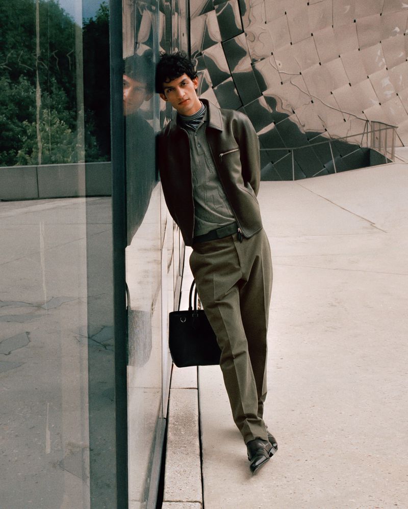 An adult male fashion model leans against a glass wall in a gray-green jacket and matching slacks, holding a black tote bag in an outdoor setting.