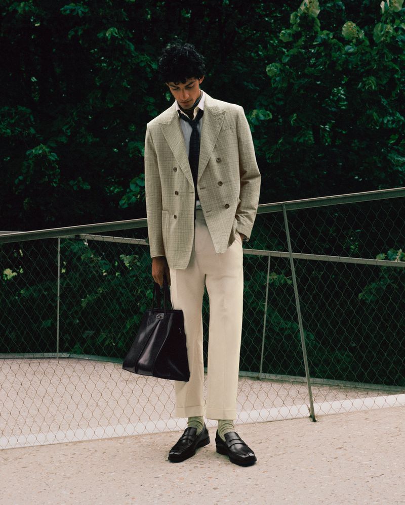 A person in a suit and tie stands outdoors holding a tote bag with a green, leafy background, creating a stylish and professional look.