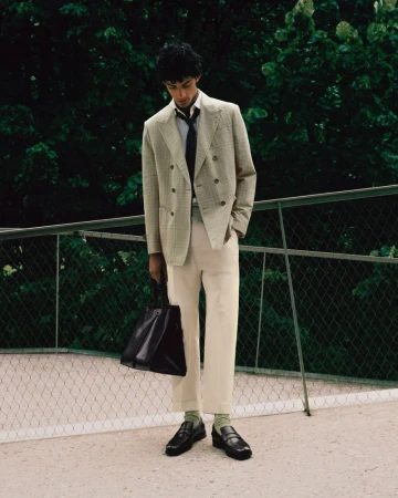 A person in a suit and tie stands outdoors holding a tote bag with a green, leafy background, creating a stylish and professional look.