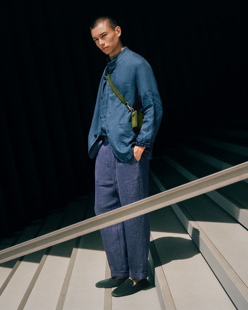 A male model poses on concrete stairs wearing a blue linen jacket, blue trousers, and black shoes with a dark background.