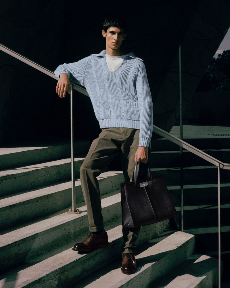 Male fashion model poses on steps in a light blue knit sweater with olive-green trousers and a black leather tote bag, outdoors.