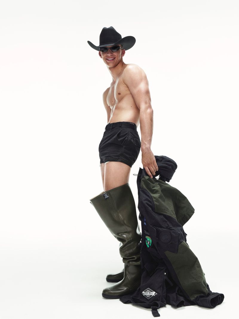A shirtless man in shorts, cowboy hat, and boots poses in a white studio, holding a jacket, showcasing an unusual combination of attire.