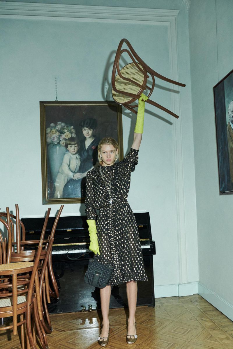A fashion photo showcases a blonde woman in a polka-dot dress, holding a chair aloft indoors with paintings, a piano, and chairs.