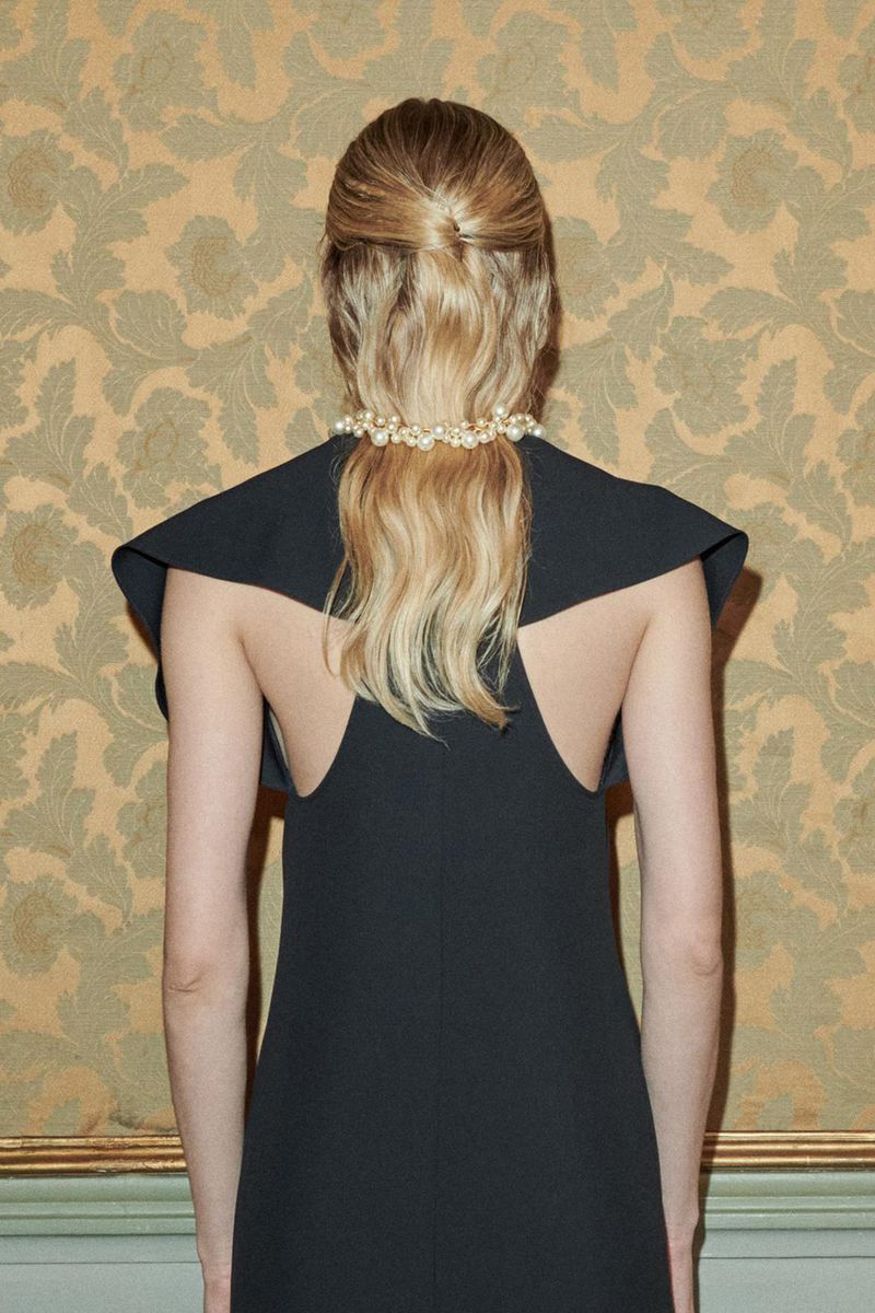 Back view of a woman in a black dress with a pearl hair accessory, standing against an ornate floral wallpaper backdrop.