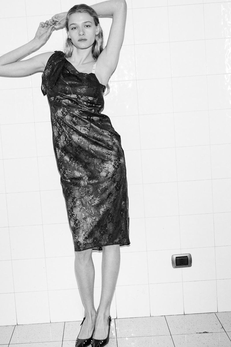 Full-length studio shot of a blonde woman in a one-shoulder black dress and black high heels against a tiled background.