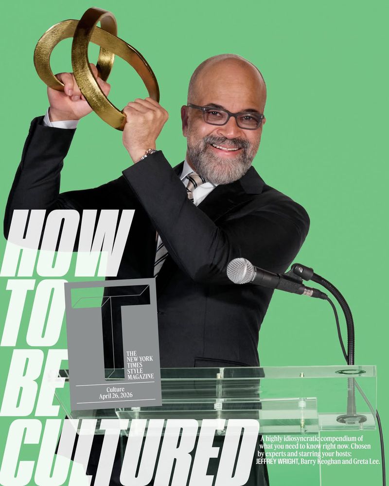 Adult man in suit behind podium raises gold rings against green backdrop. Magazine cover with headline, How to Be Cultured visible.