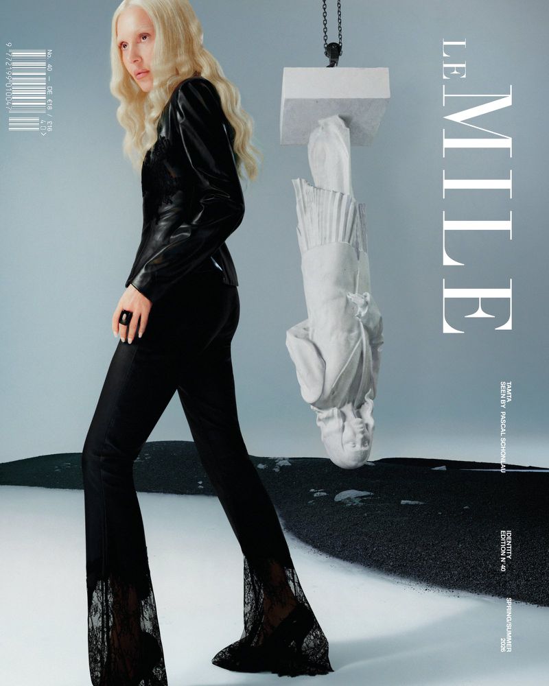 Fashion editorial shot of a fair-skinned woman in black pants with lace, a top, and platinum hair, posed with an art sculpture.