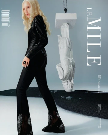 Fashion editorial shot of a fair-skinned woman in black pants with lace, a top, and platinum hair, posed with an art sculpture.