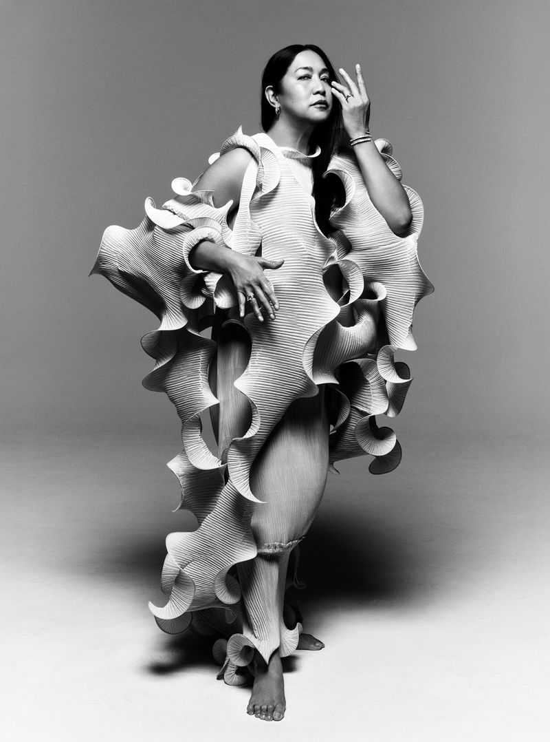 Portrait of an Asian woman in a full-length shot, wearing a textured avant-garde dress against a grey backdrop in a photo studio.