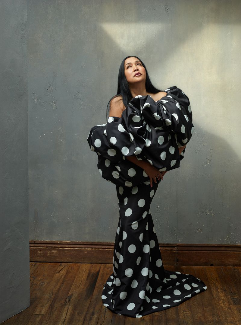 Woman in a black and white polka dot dress looking up, standing against a textured grey wall with a wood floor and natural lighting.