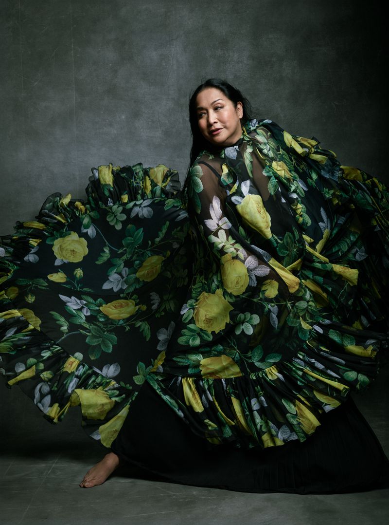 An Asian woman posing in a floral gown in a studio. She is barefoot and smiling, and the gown has a large ruffled skirt.