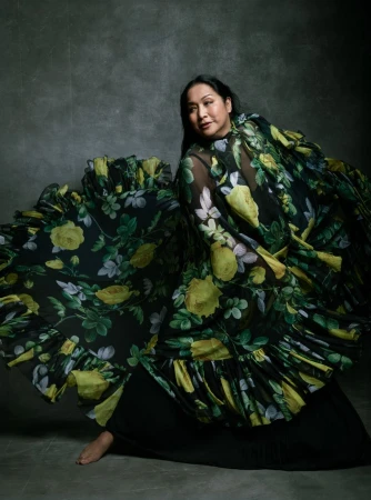 An Asian woman posing in a floral gown in a studio. She is barefoot and smiling, and the gown has a large ruffled skirt.
