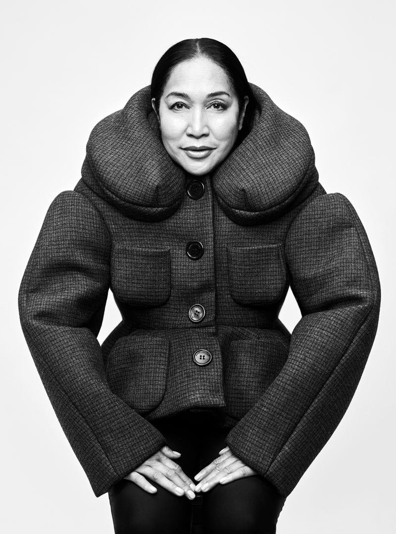 Black and white portrait of an adult woman wearing a large, structured tweed jacket against a plain background, hands clasped in front.