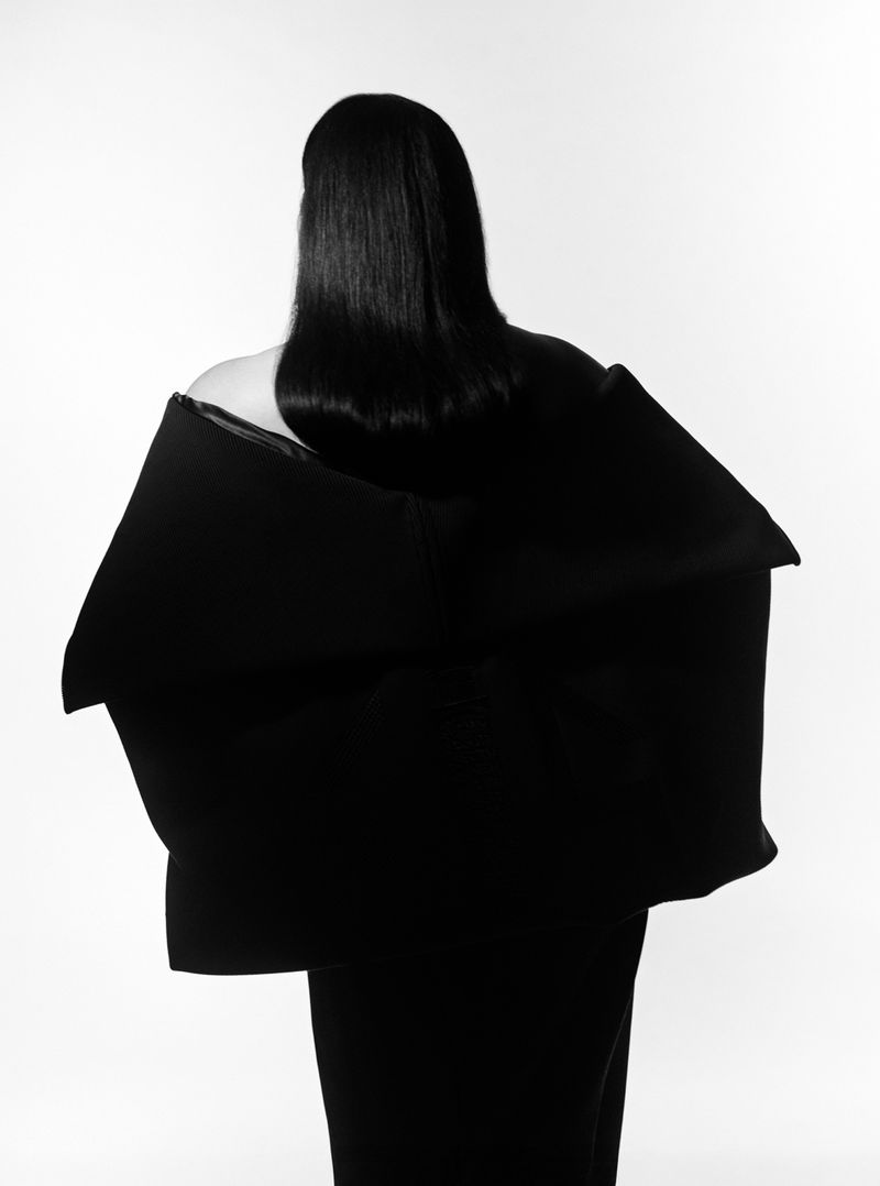 Back view of a person with long dark hair wearing a large black shawl or jacket against a white background in a studio setting.