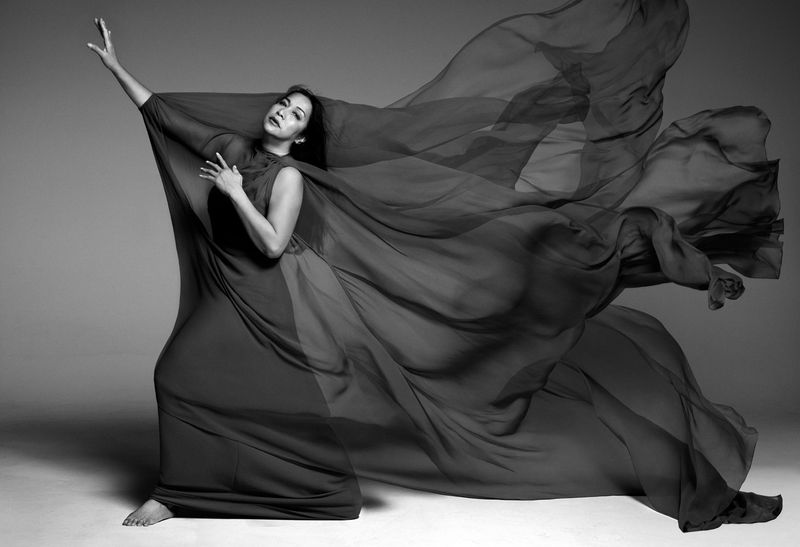 Elegant monochrome portrait of a woman in a flowing dress with arms raised against a simple backdrop, emphasizing movement and grace.