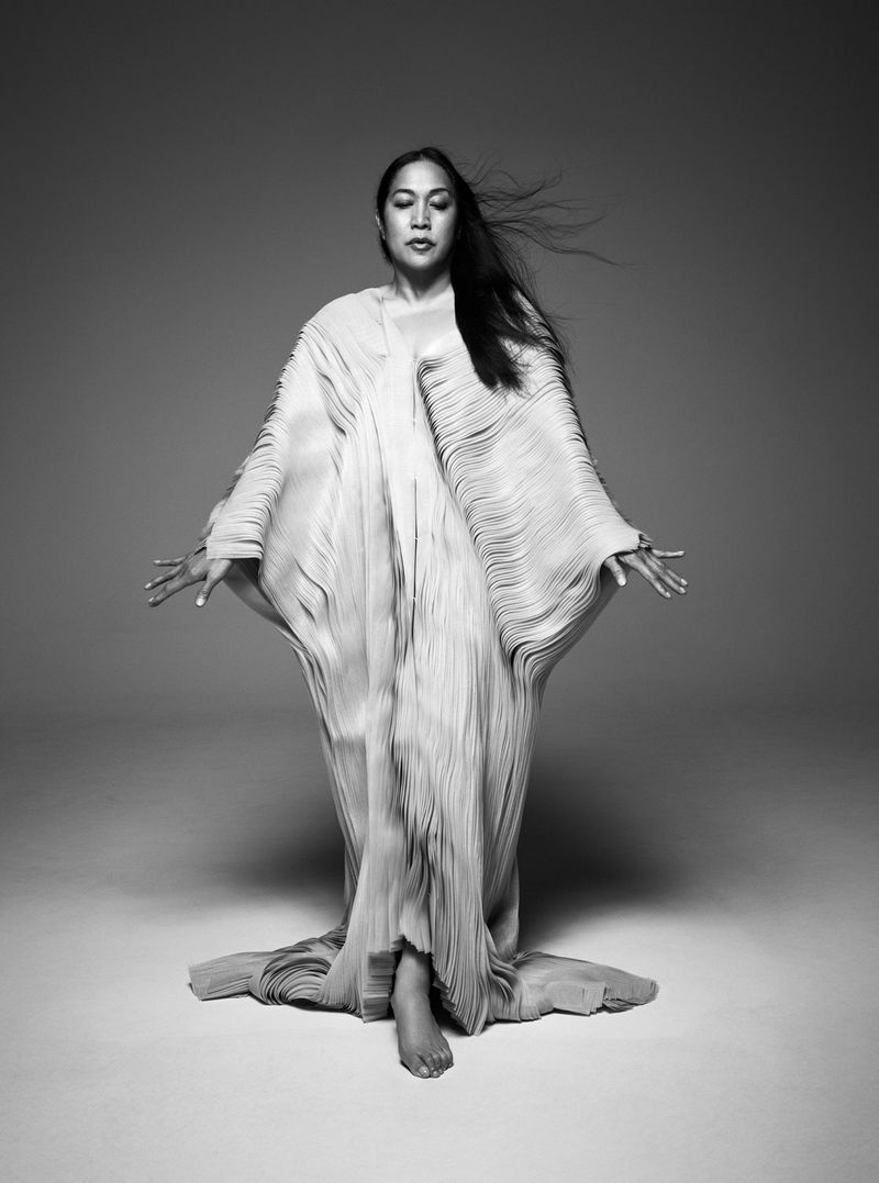 Black and white portrait of a woman wearing a flowing, textured gown. She is standing barefoot with arms outstretched in a studio setting.