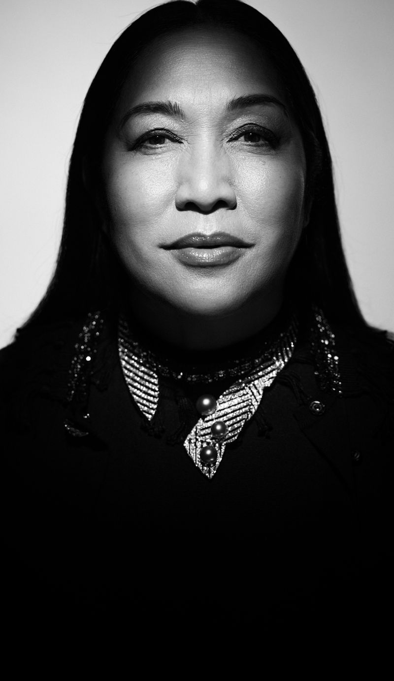 Portrait of an adult female with long dark hair, wearing a textured dark blazer and a decorative collar, against a white background.